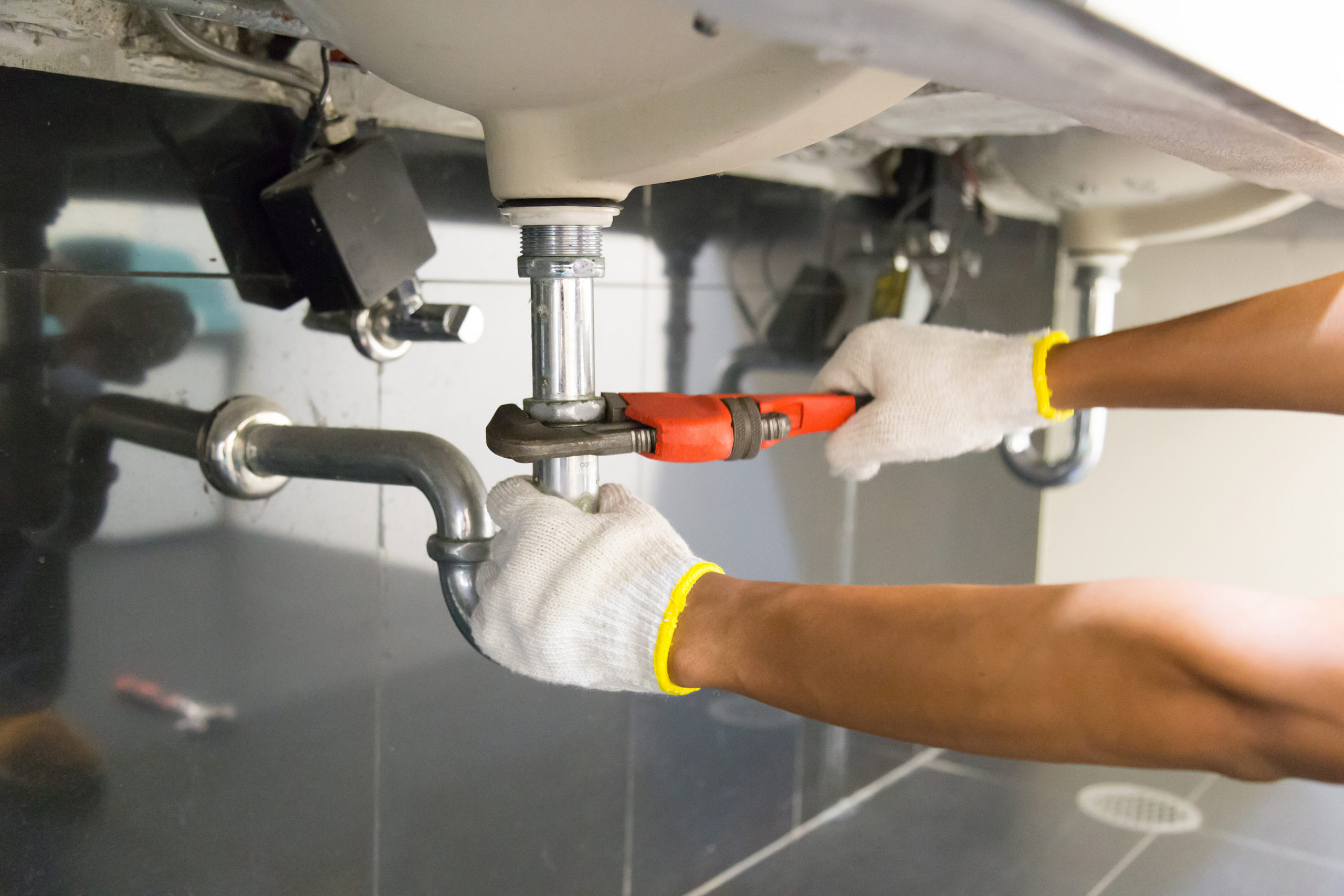 Plumber fixing white sink pipe with adjustable wrench. Plumber inspecting pipes under sink for regular home plumbing maintenance.